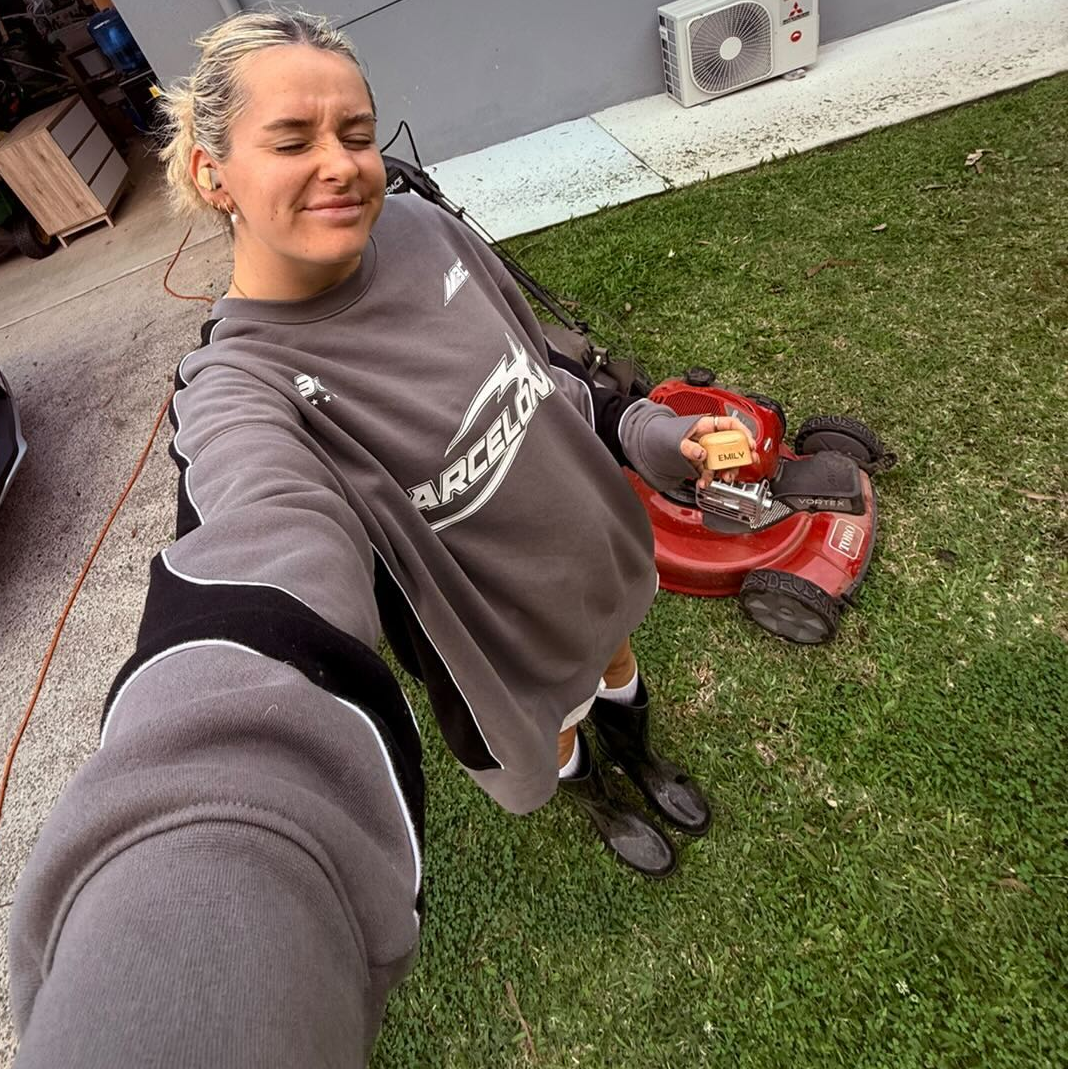 Emily taking a selfie by her push mower holding a PureBuds Pro bamboo charging case and wearing Her PureBuds Pro bamboo wireless earbuds
