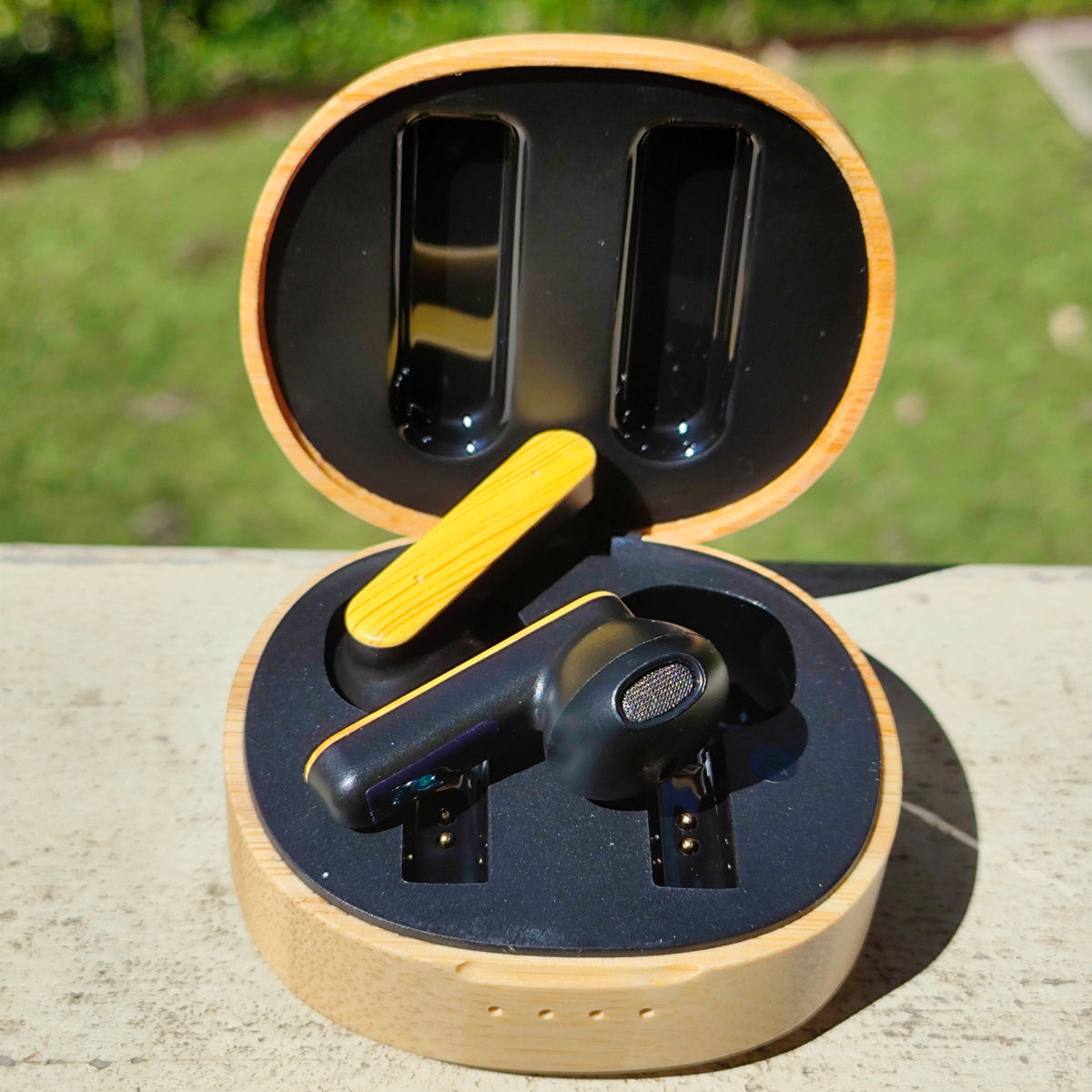 We Pure PurePods open-fit earbuds in a natural bamboo charging case with black accents, set outdoors in sunlight.